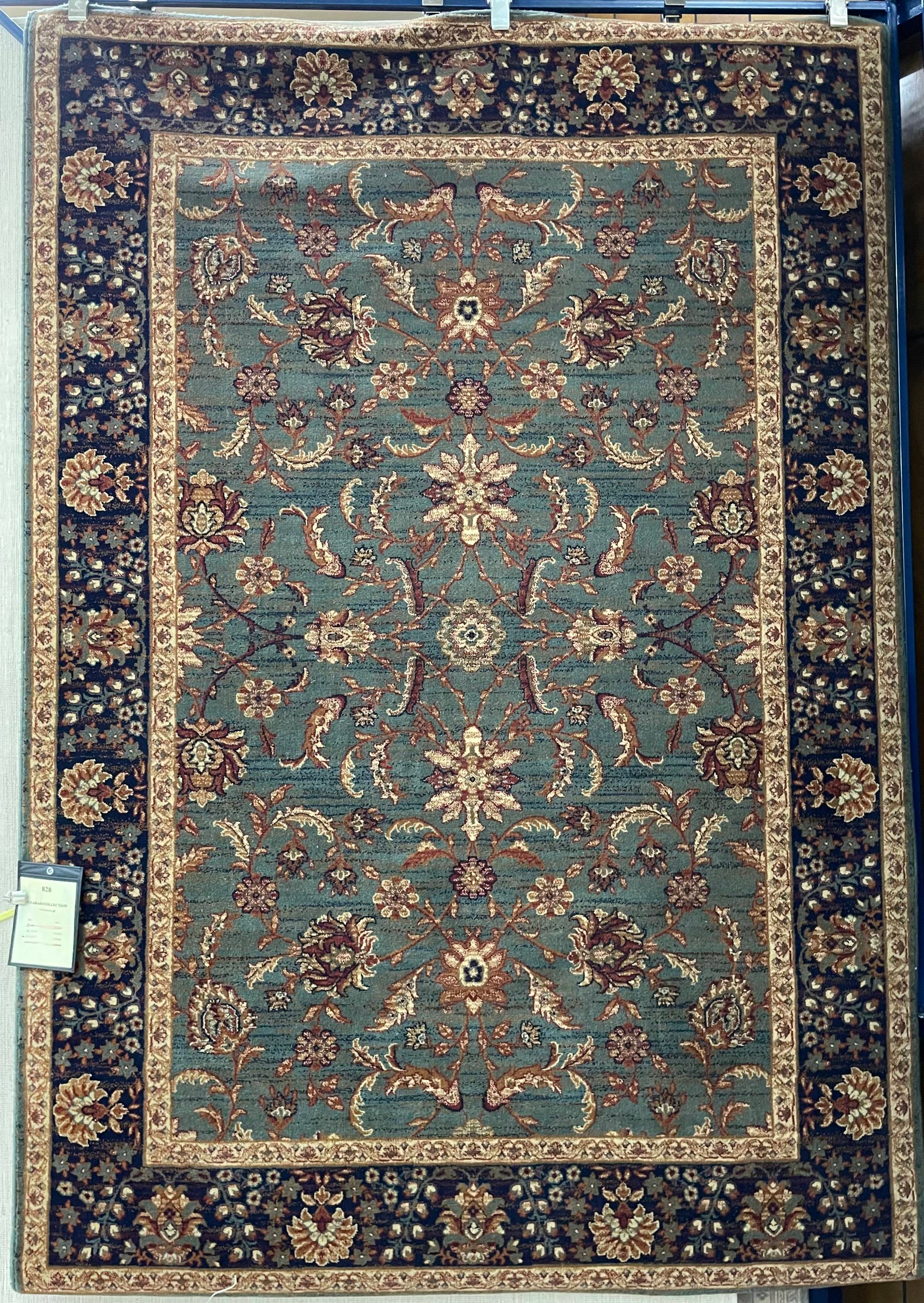 The image depicts a rectangular rug with an intricate design. The symmetrical pattern features a central medallion surrounded by floral motifs and curvilinear vine patterns. The color palette predominantly includes shades of beige, brown, and teal. A layered border frames the central field, and a small white tag is visible in the bottom left corner.