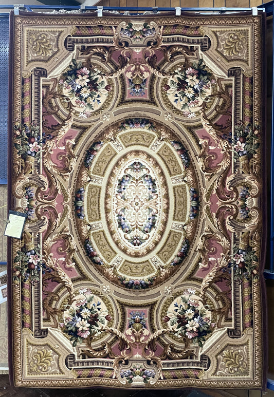 The image depicts a rectangular rug with an intricate design. The symmetrical pattern features a central oval medallion surrounded by intricate floral patterns and scrollwork. The color palette includes shades of beige, pink, and green on a dark background. The rug is displayed hanging against a vertical surface with tags visible on the lower left side, indicating it may be for sale in a retail setting.