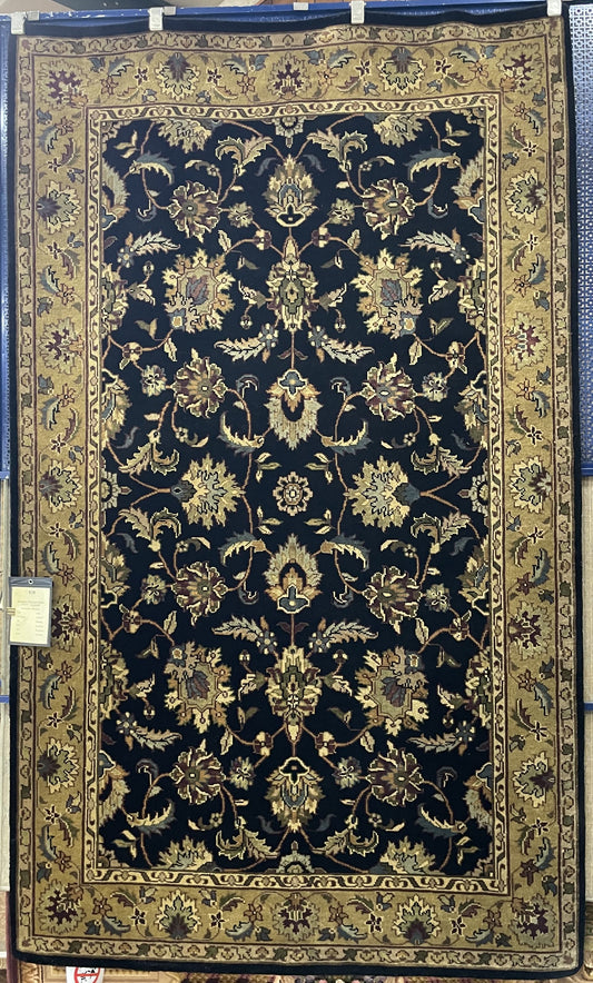 The rug in the image features a dark background—possibly black or deep navy—with an intricate pattern of floral and vine motifs. The colors appear to be gold, tan, and lighter shades of blue. The design is symmetrical and ornate, with a densely filled central field and a complementary border.